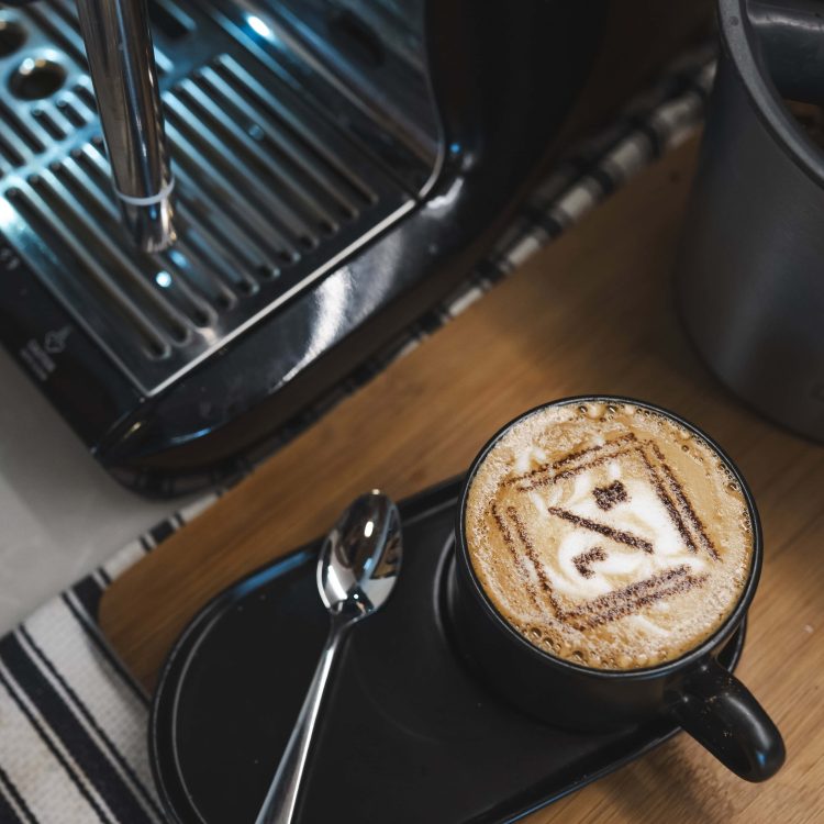 Frothy coffee with latte art on a wooden table, coffee machine in background.